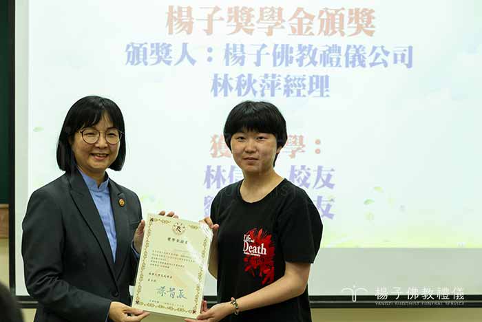 在南華大學的教室裡，投影幕上顯示著「楊子獎學金頒獎」，楊子佛教禮儀代表林秋萍經理與獲獎碩士班同學劉育瑄溫馨合照。｜楊子佛教禮儀公司